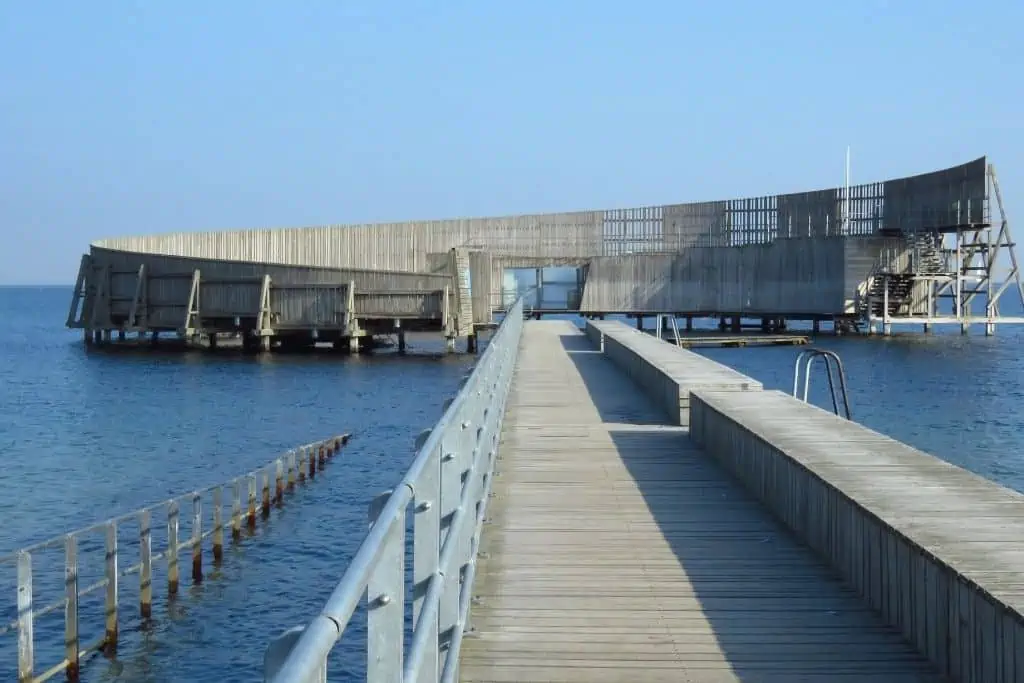 Kastrup海浴，一个木质平台延伸到水面上的圆形建筑，保护游泳者和日光浴者免受风的伤害，同时创造了一个阳光陷阱。